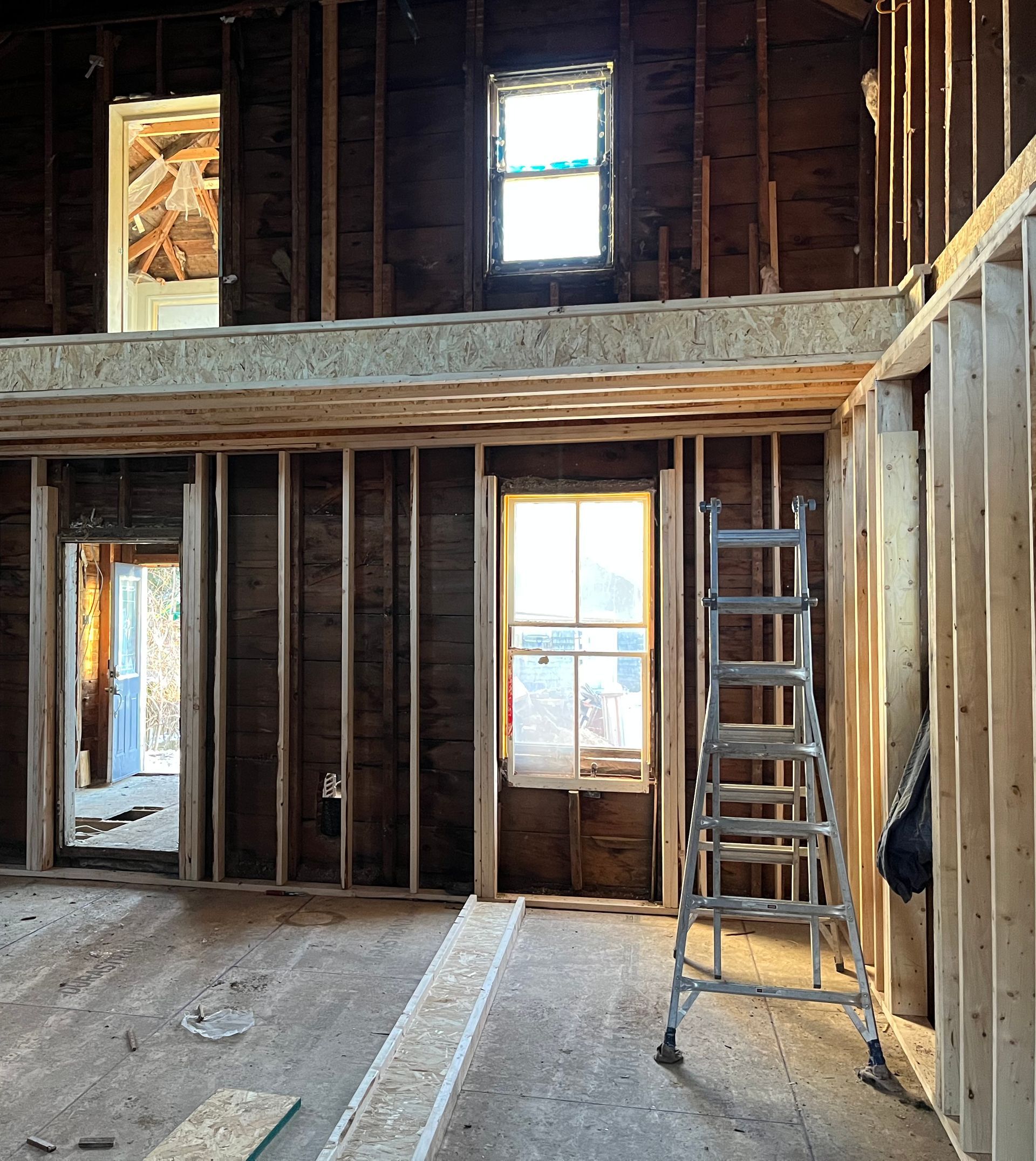 Interior of a house under renovation. Bare wood framing, windows, and a ladder are visible.