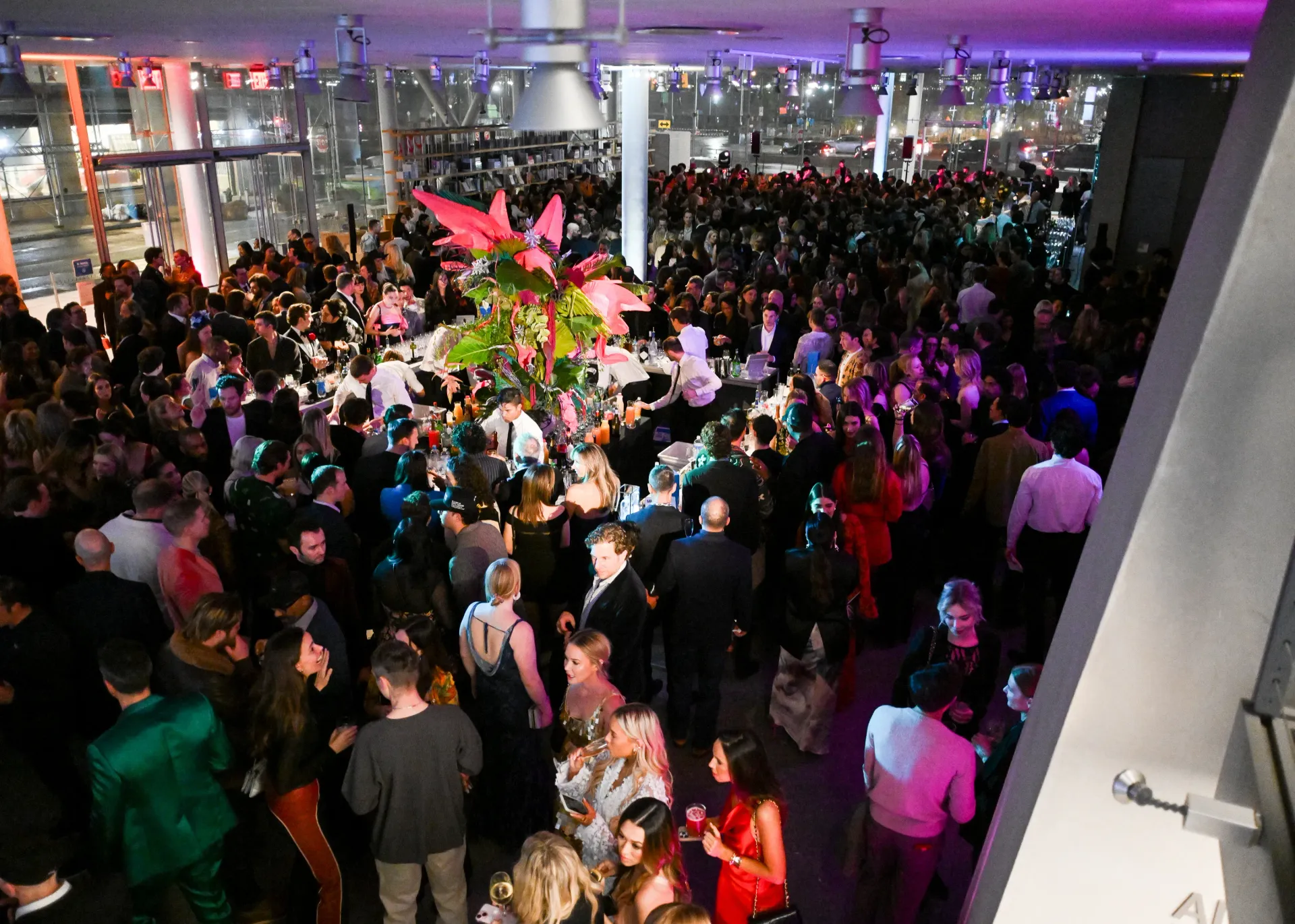 Crowded indoor reception with guests mingling under purple lighting and a large floral centerpiece