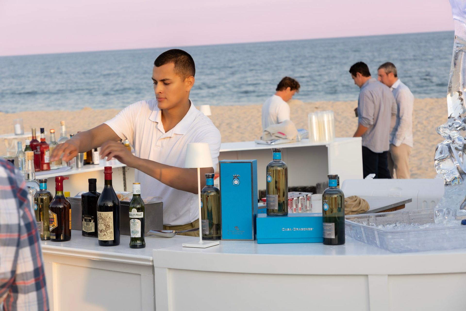Bartender serving drinks at an outdoor beach bar with bottles and ice under a pink sky