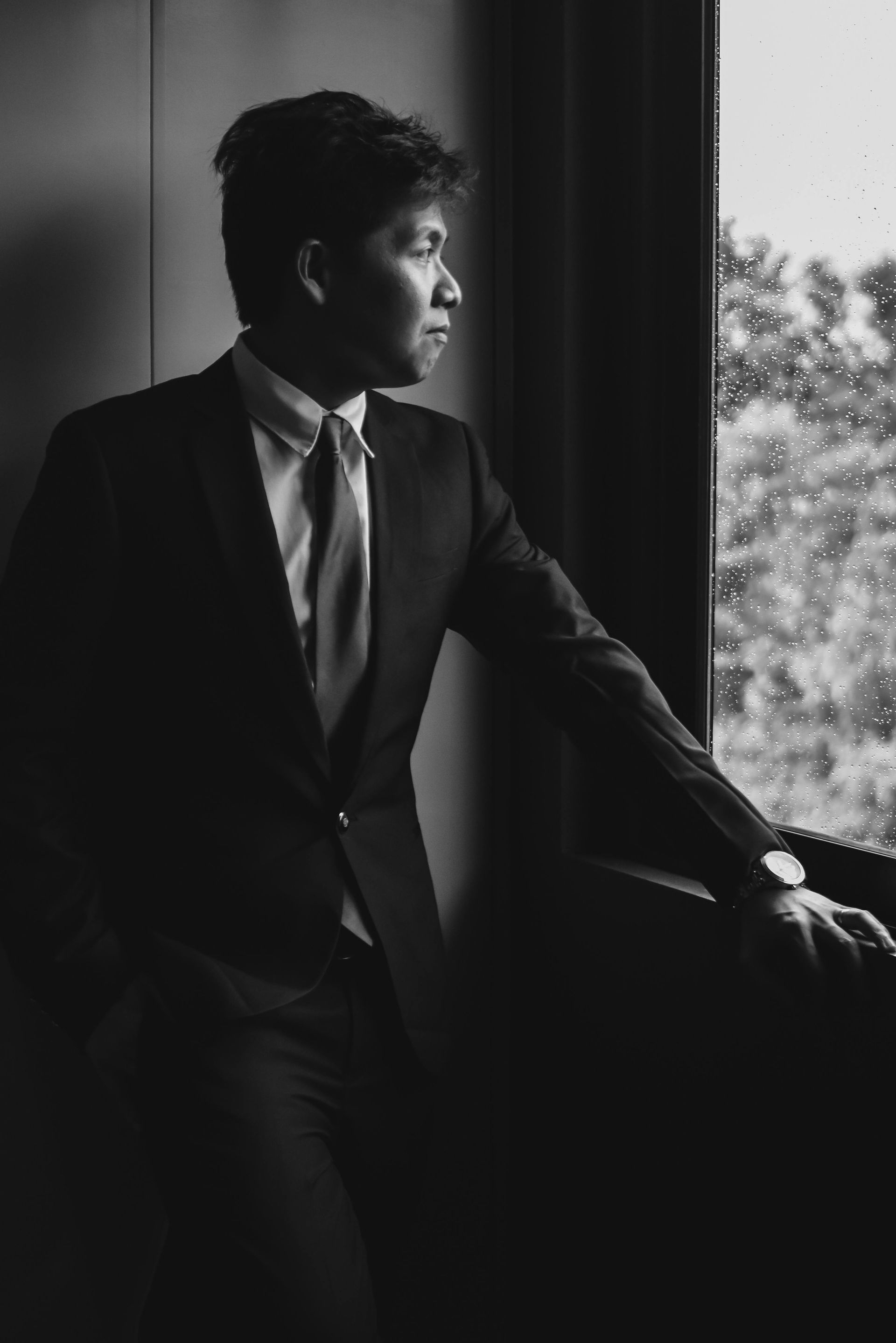 Man in a dark suit gazing out a window, standing in soft black-and-white light