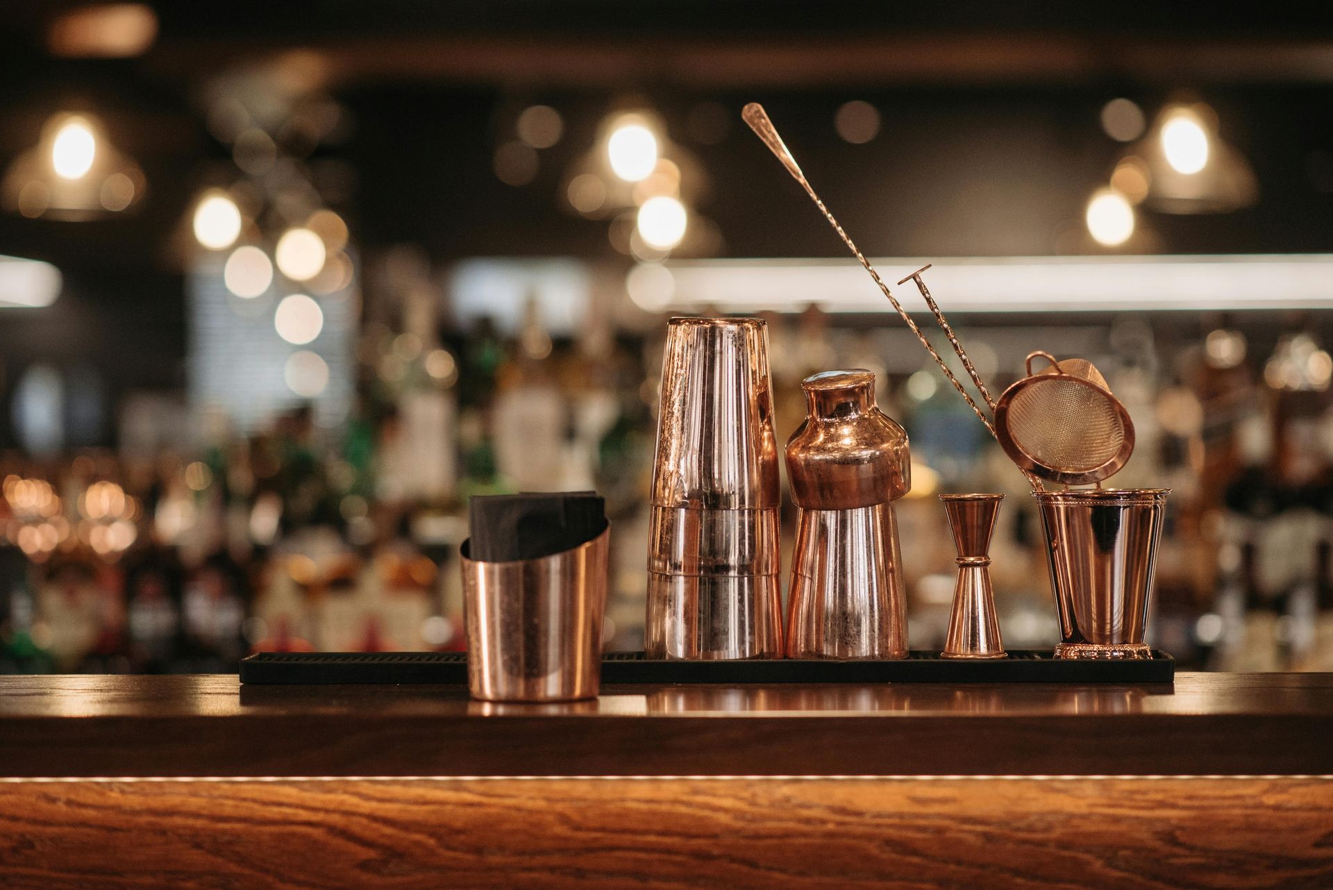 Bar tools on a wooden counter in a warmly lit cocktail bar, with shakers and strainer in the foreground
