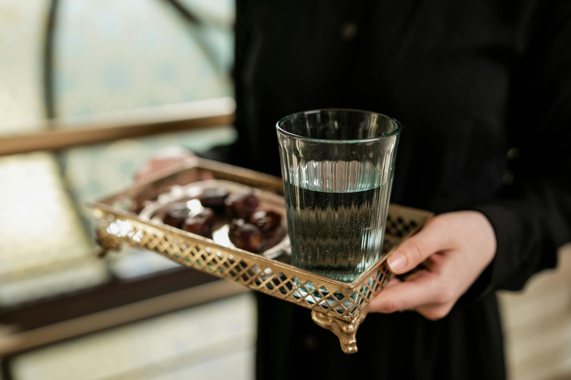 Person holding a gold tray with a glass of water and chocolates near a window