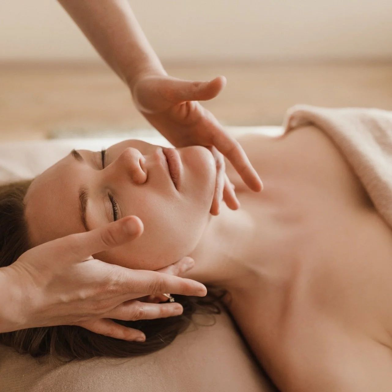 Woman receiving facial massage, hands on face and neck, eyes closed, in a spa-like setting.