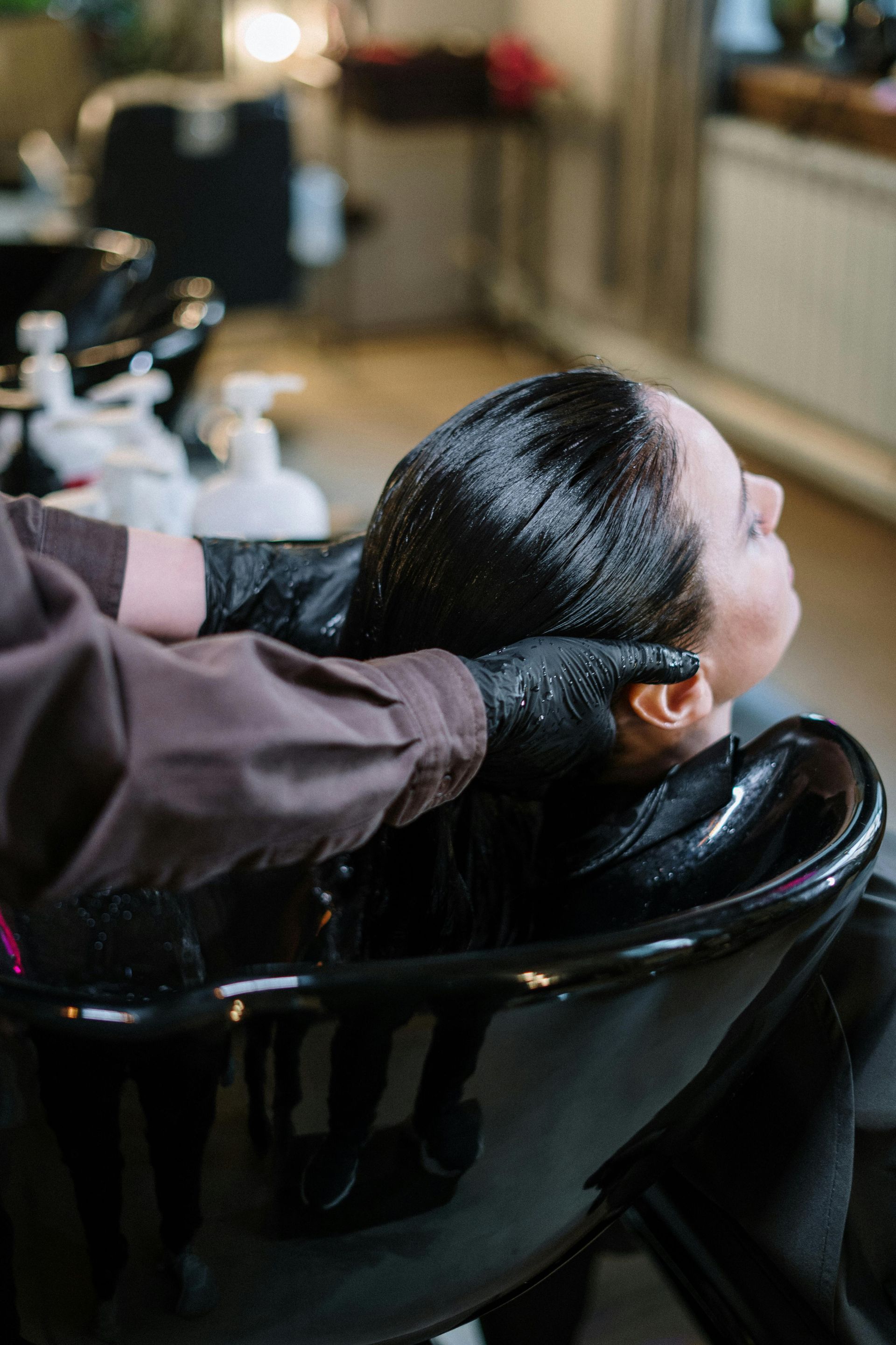 Hairdresser washing a client's dark hair over a black sink. They are wearing black gloves.