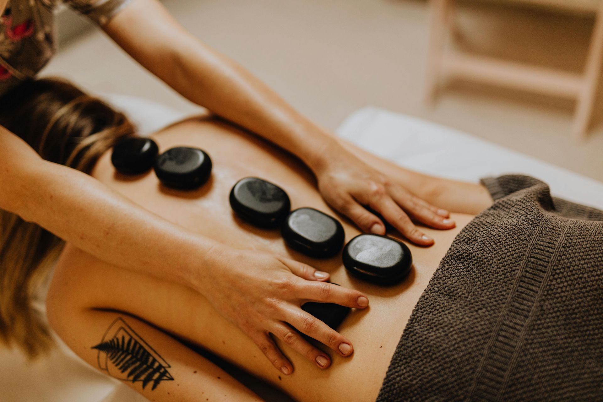 Woman receiving a hot stone massage on her back, with stones placed and masseuse's hands on her.