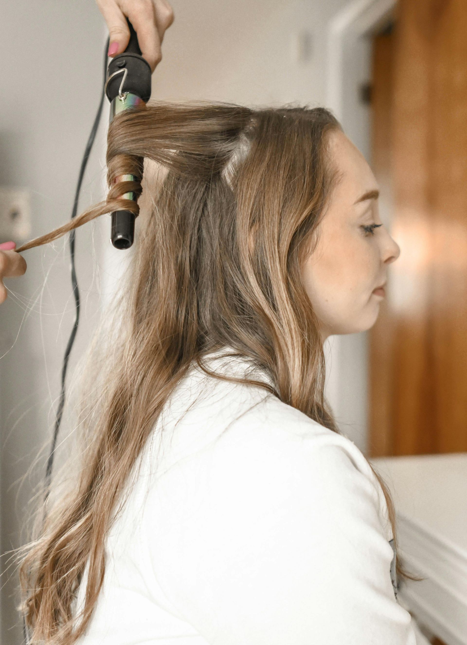 Woman getting her hair curled with a curling iron, white top, side profile view.