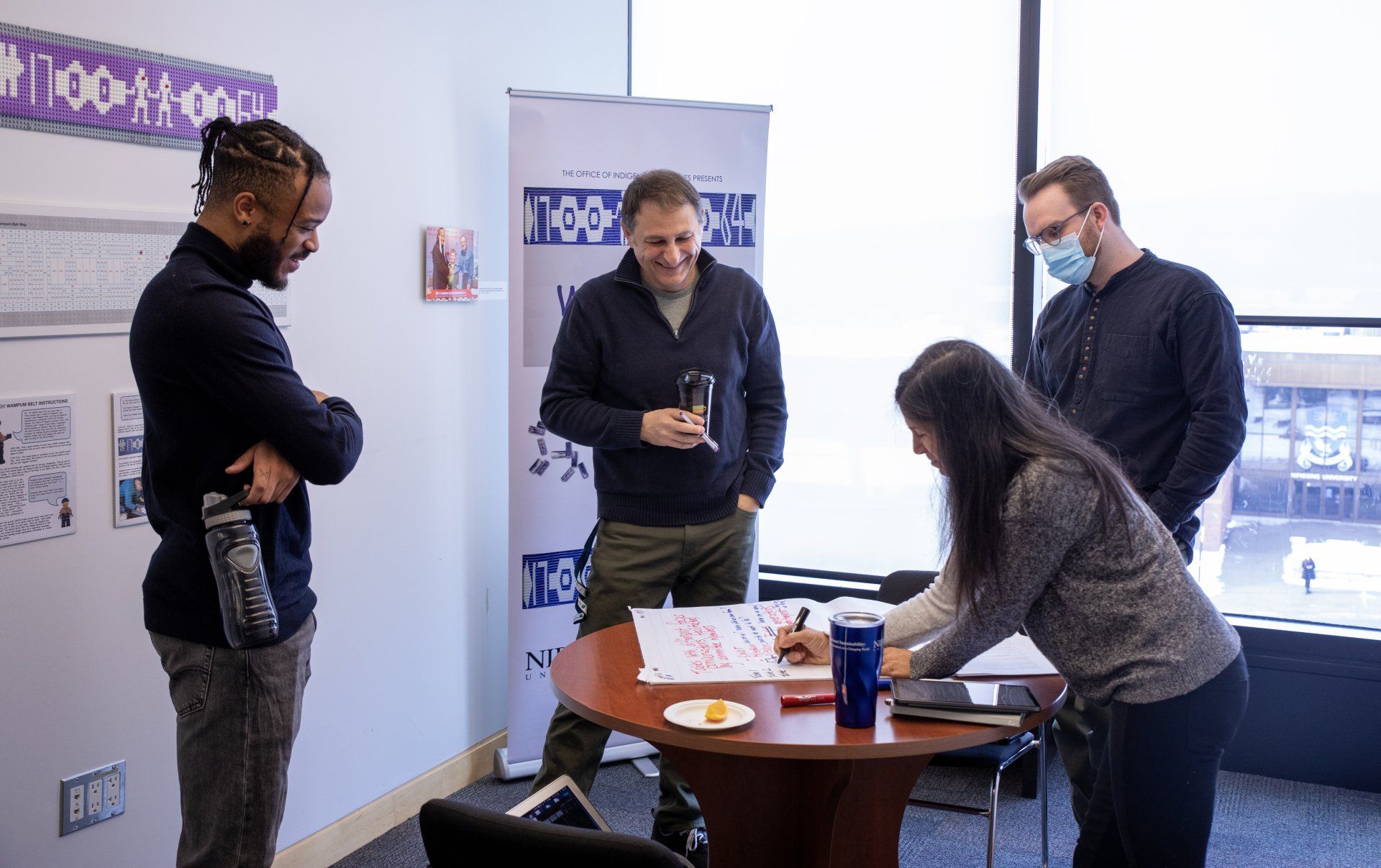 Four people in a modern office, collaborating around a table. One person signs a paper; others observe and converse.