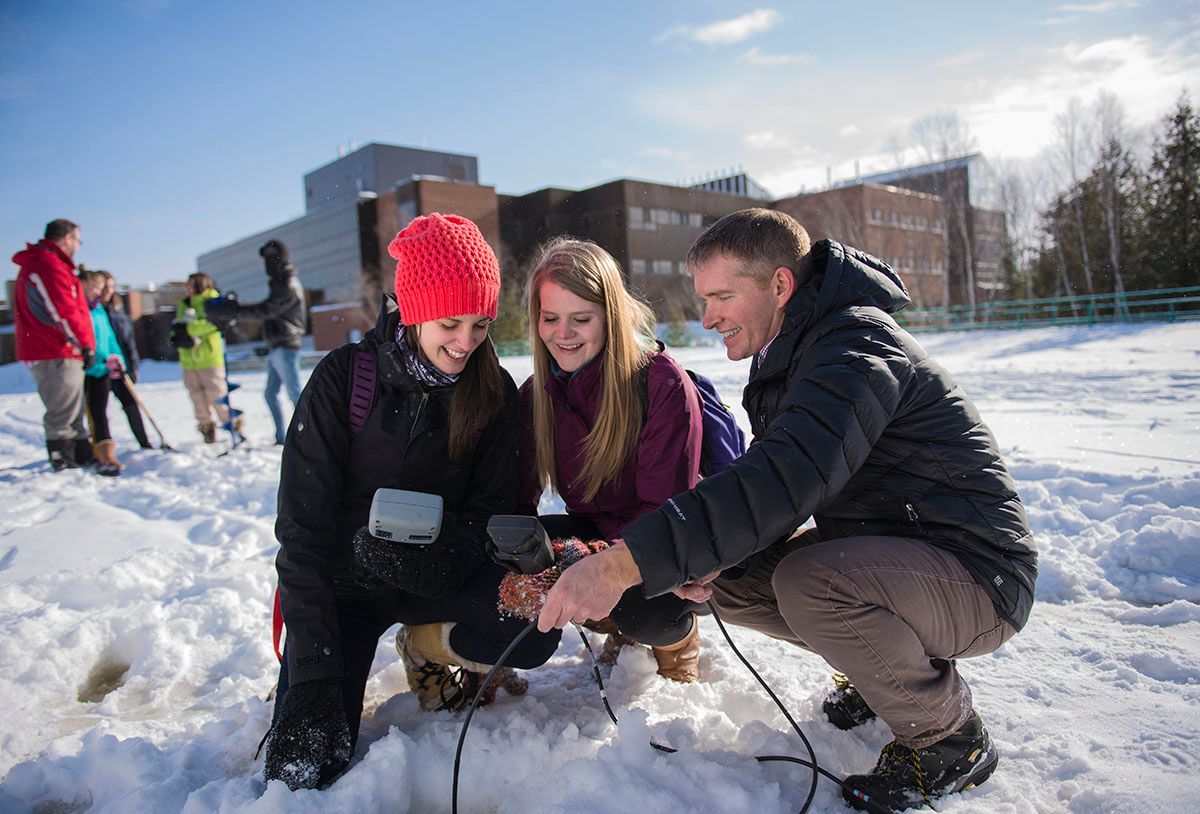 Three people examining equipment in snow, Nipissing University in background.