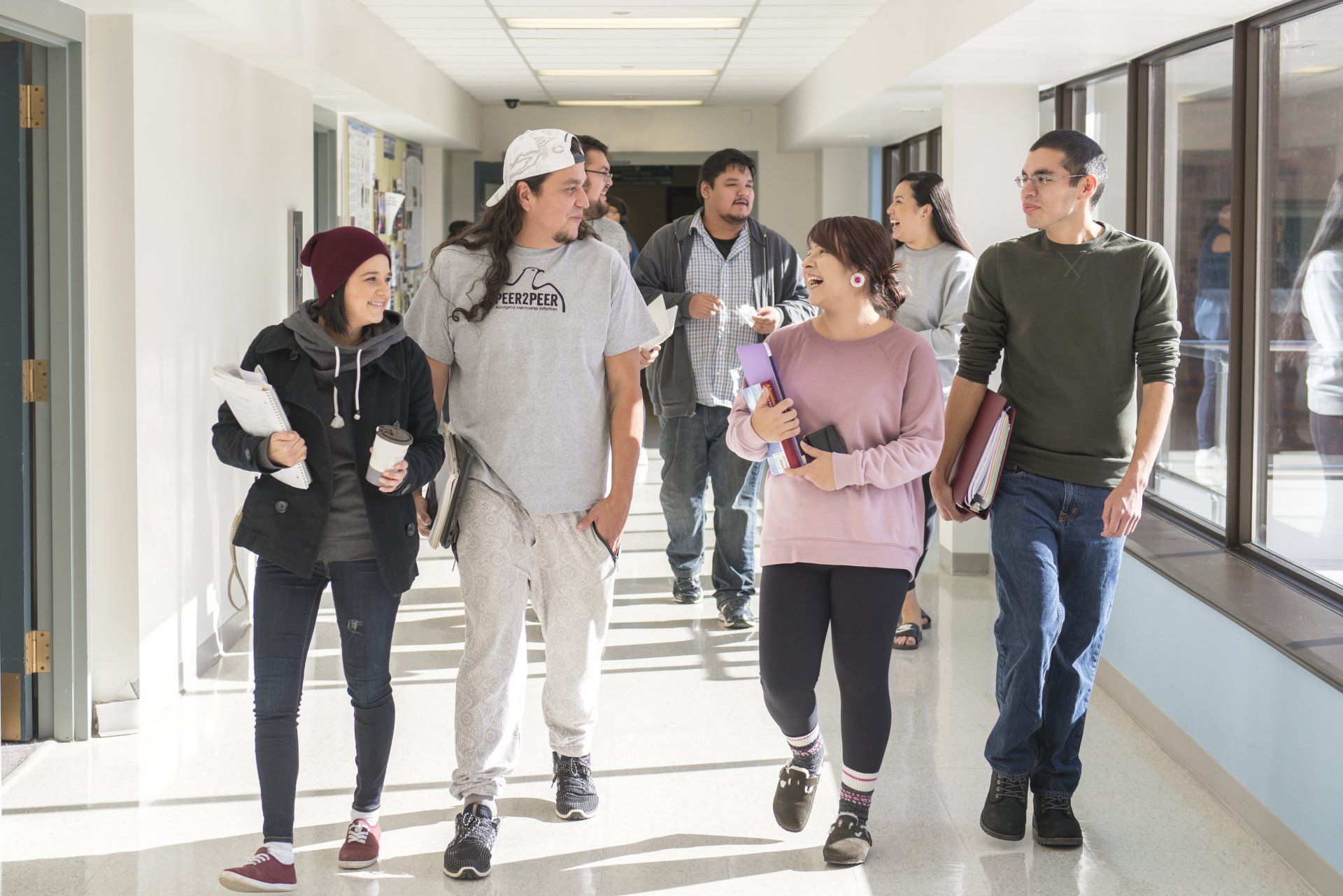 Students walking and talking in a brightly lit hallway, some carrying books and coffee.