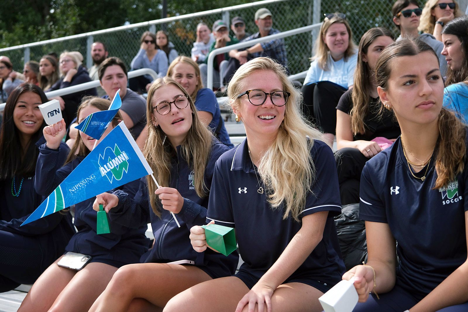 Spectators at a sporting event; women cheering, holding flags and cups, wearing blue athletic shirts.