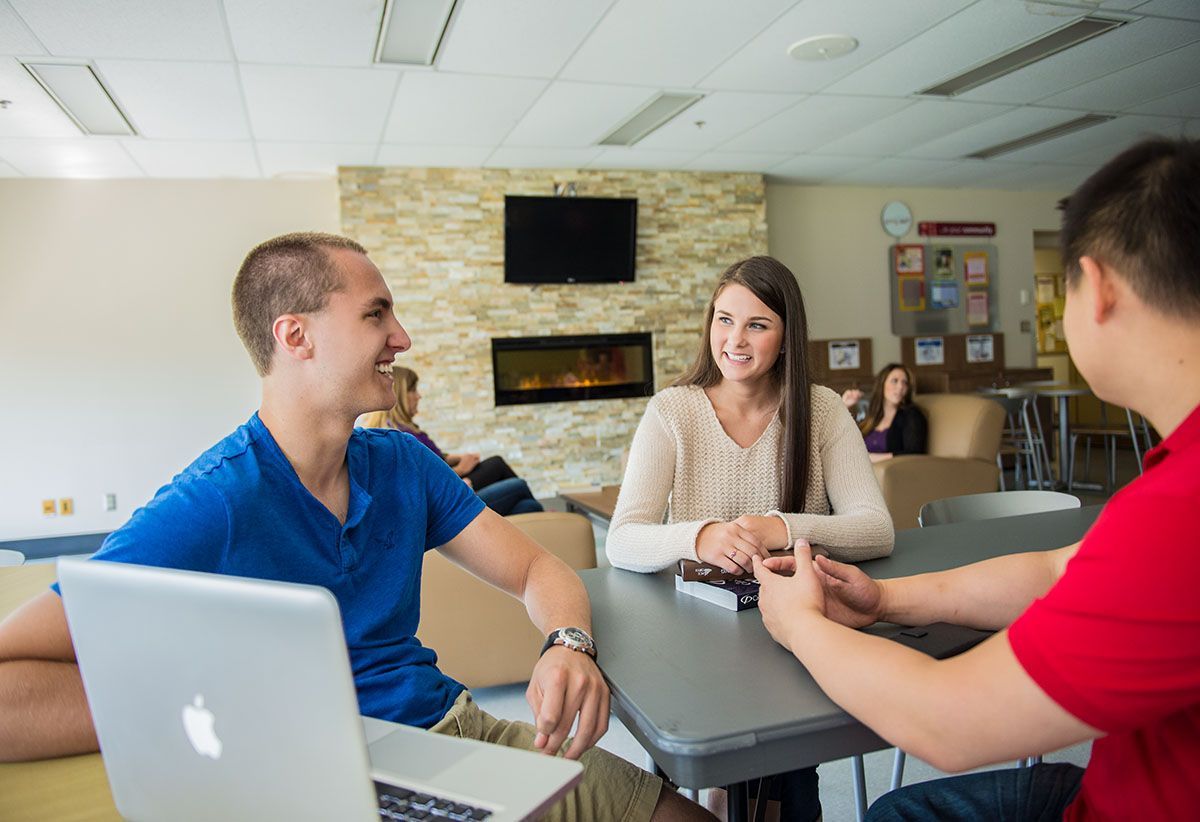 Three students sit around a table in a lounge, smiling and talking. One has a laptop open.