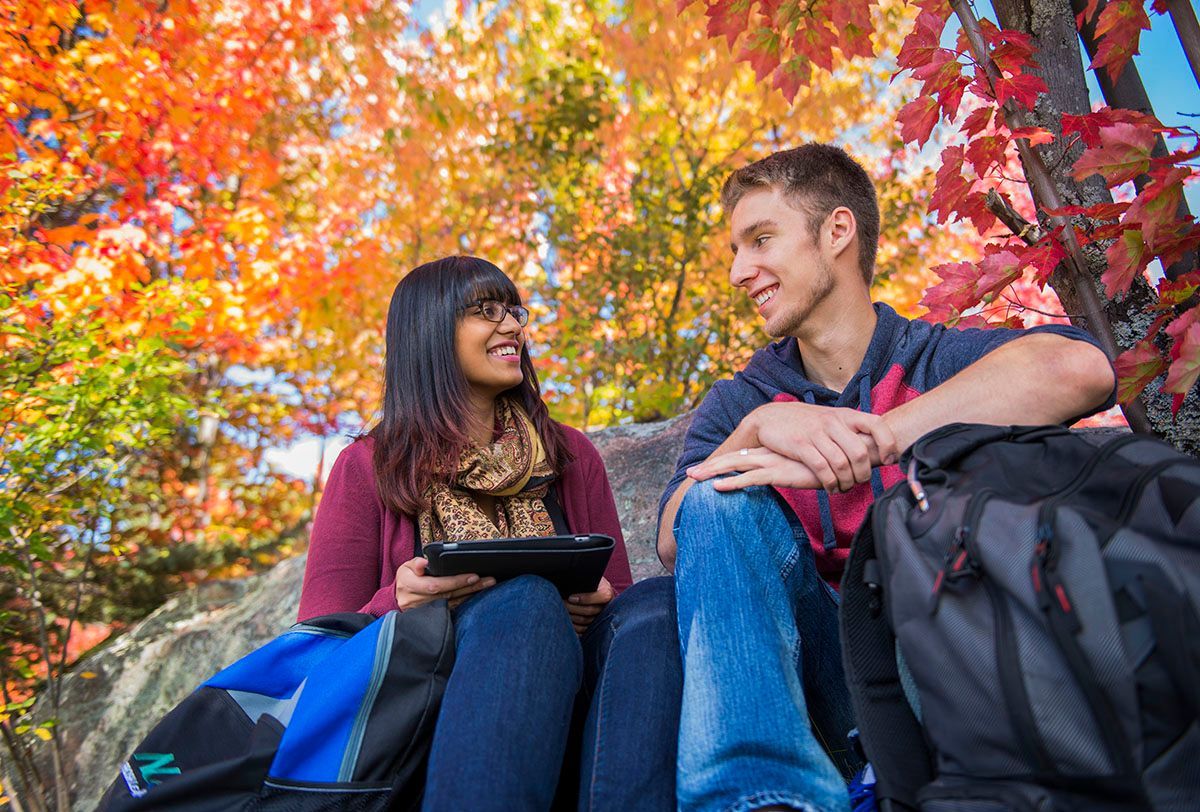 Two students seated outdoors, smiling, looking at each other. Surrounded by colorful autumn foliage.