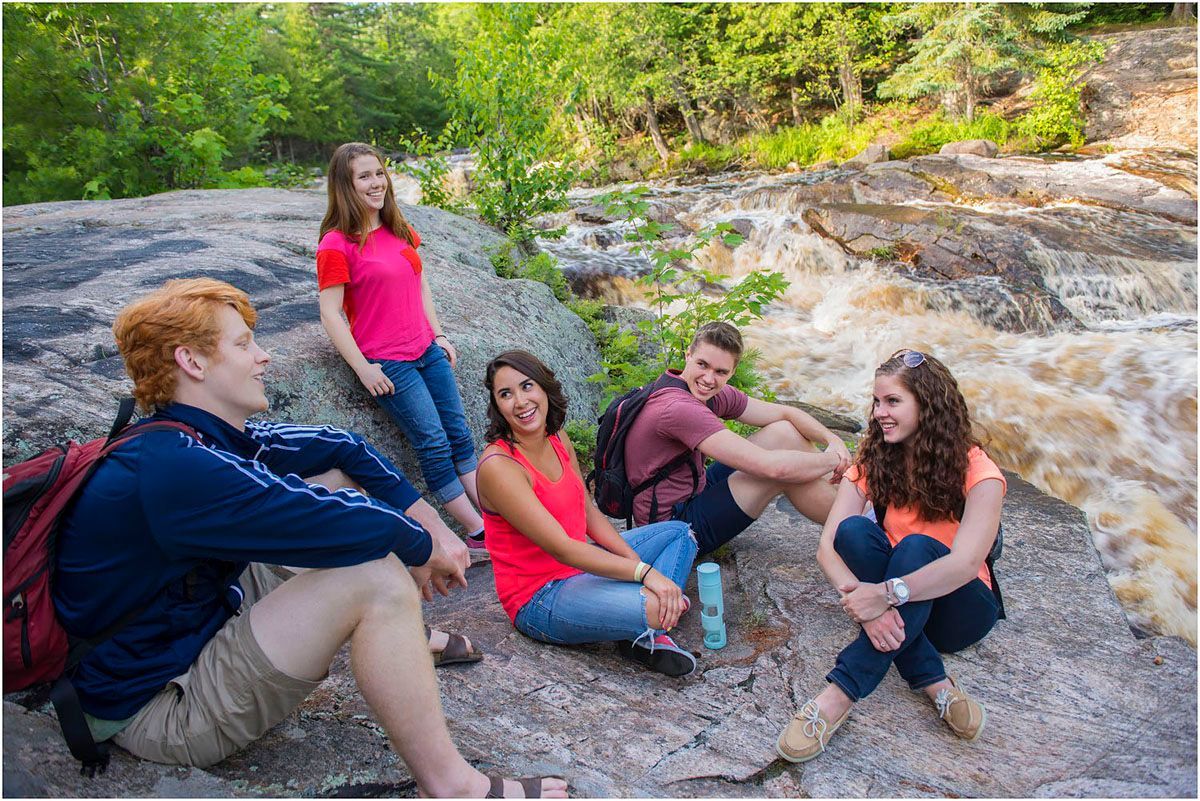 Five students sitting on a rock by a rushing river, smiling. Summer day, wearing casual clothes.