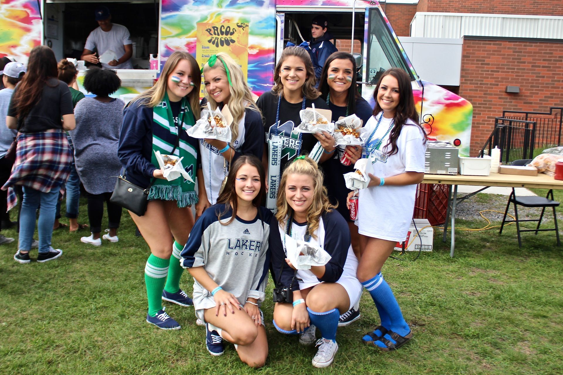 Group of smiling young women near a food truck, some holding food; outdoors.