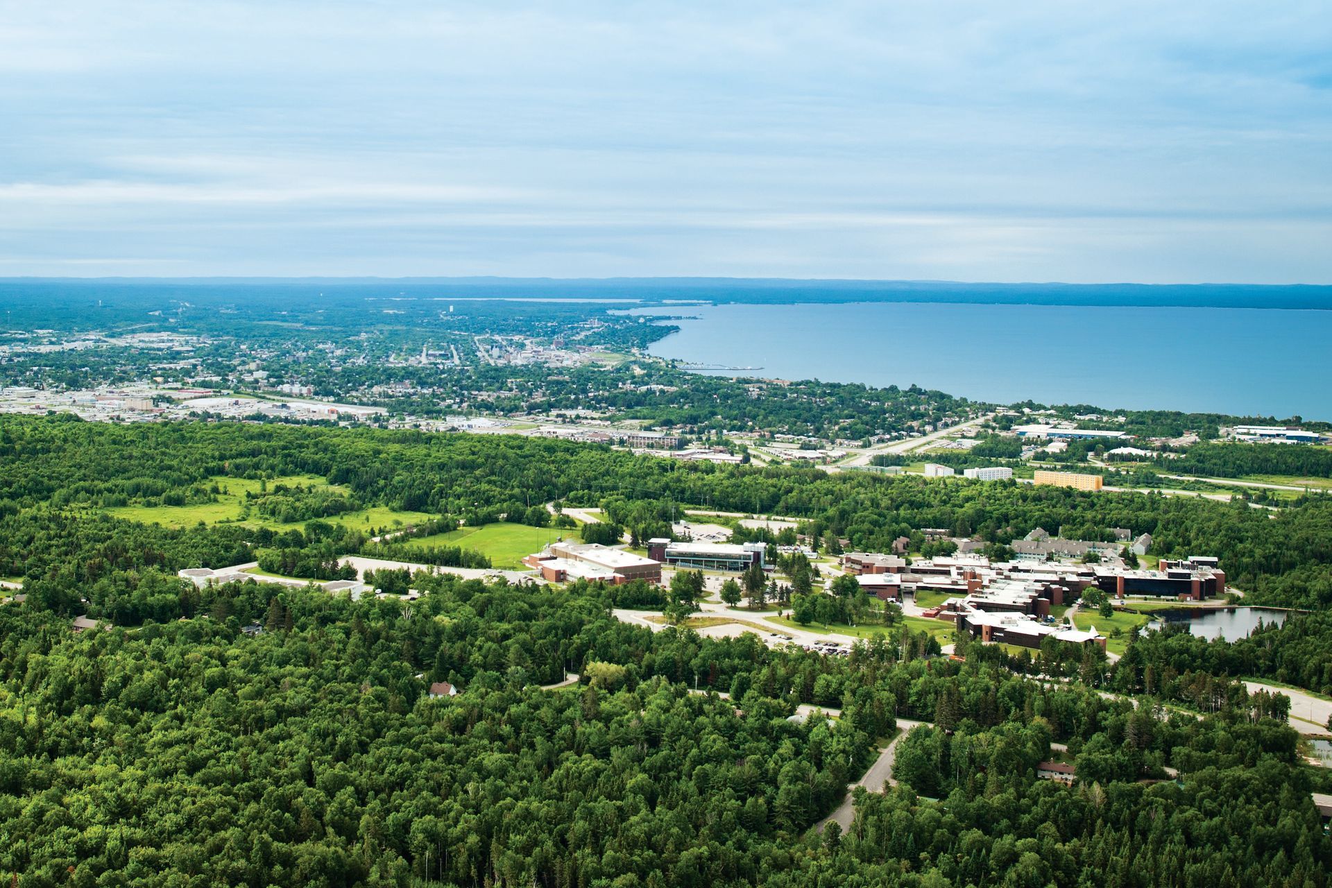 Aerial view of a college campus nestled near a lush forest and waterfront, with a cloudy sky.