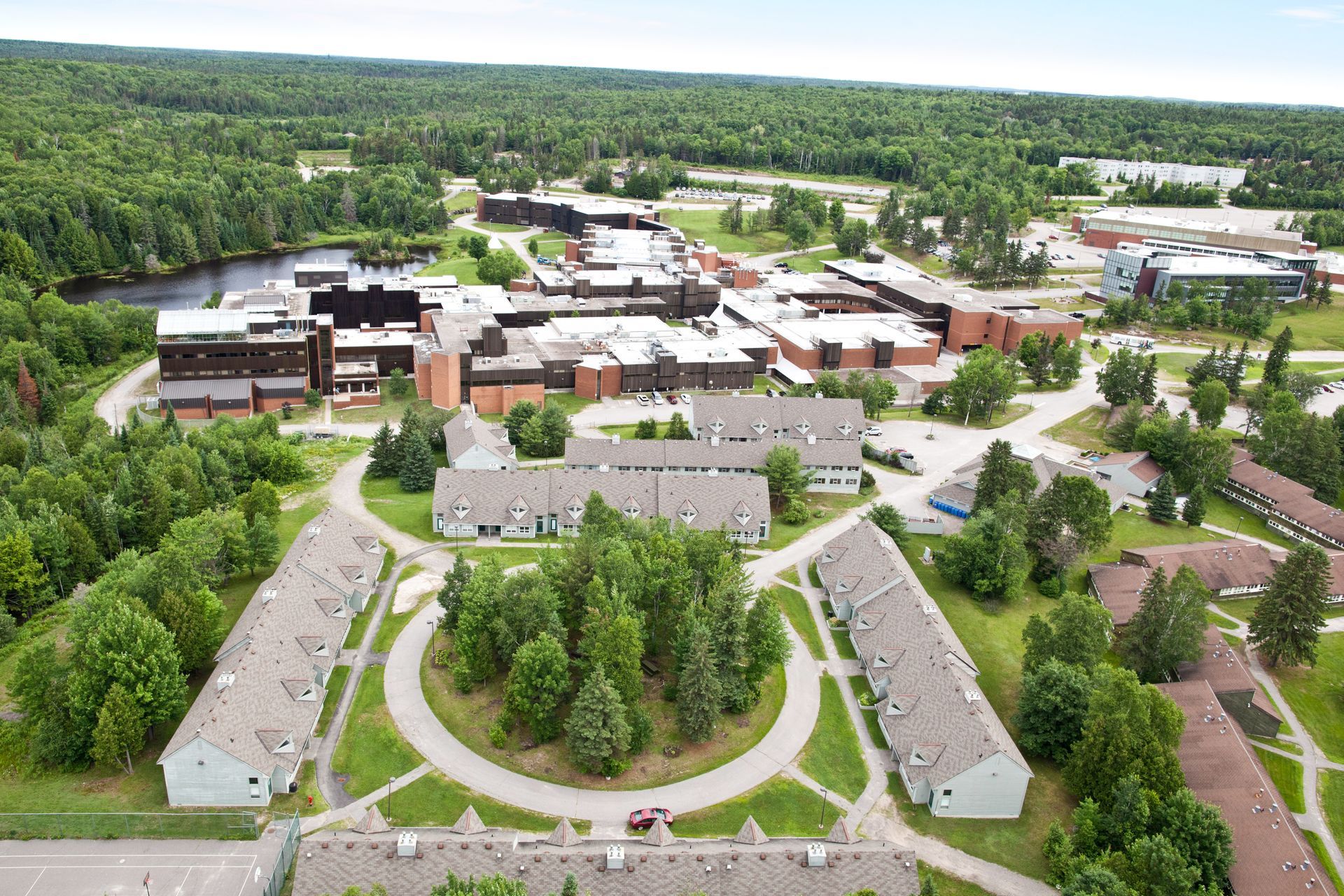 Aerial view of a Nipissing University campus surrounded by trees, with multiple buildings, green grass, and a water body.