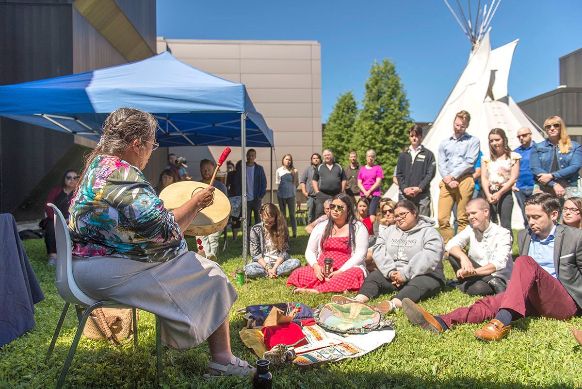 Woman drumming, surrounded by people in a circle, outdoors near a tipi and building.