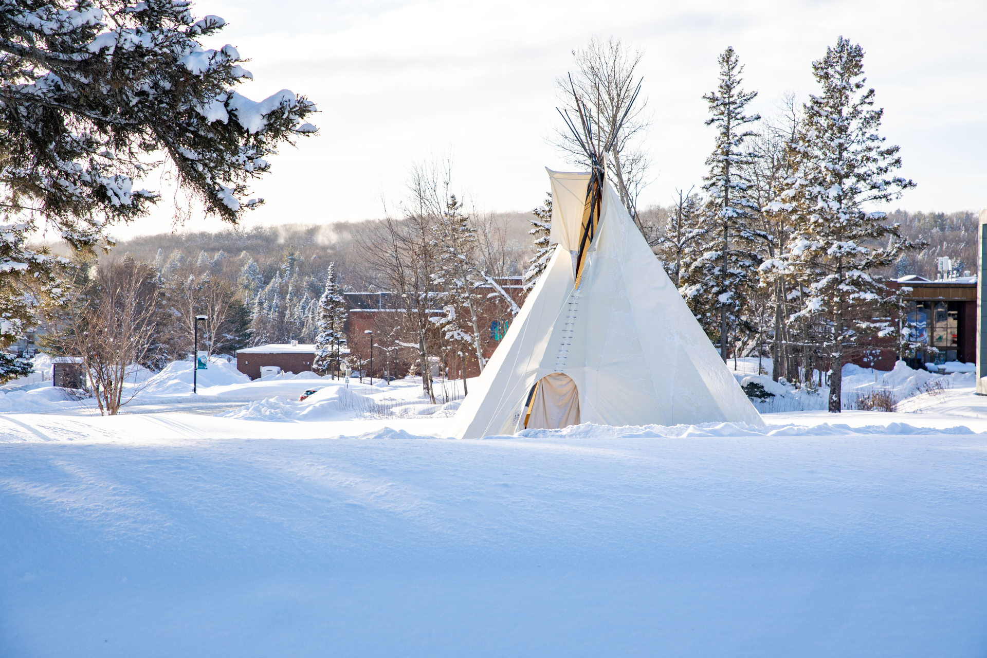 Snow-covered field with a white tipi, trees, and buildings in the background under a bright sky.