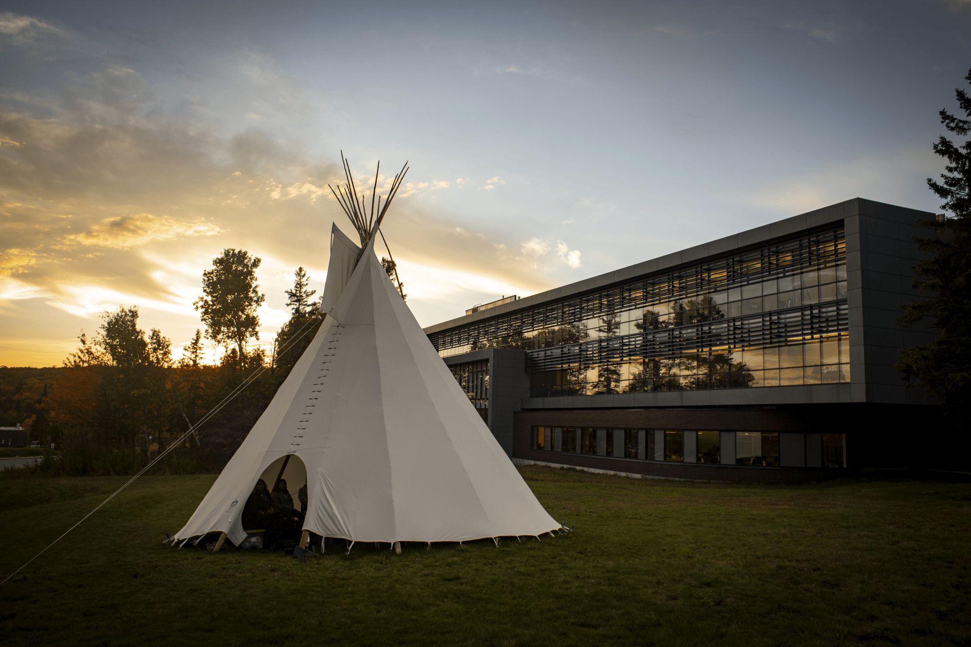 Tipi in front of a modern building on a grassy lawn, sunset sky in the background.
