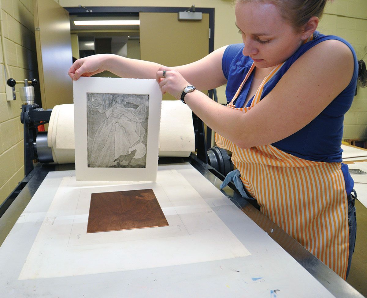 Woman in apron printing artwork using a press; she holds up the print, looking down.