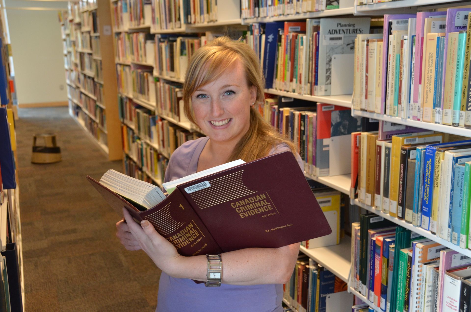 Woman in a library smiles, holding an open maroon book; bookshelves fill the background.
