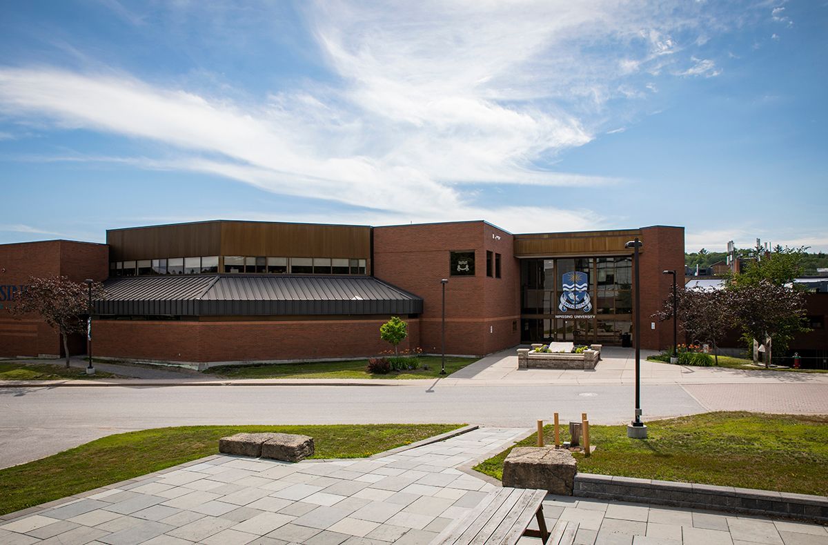 Nipissing University building under a blue sky, with a walkway and green lawn in the foreground.