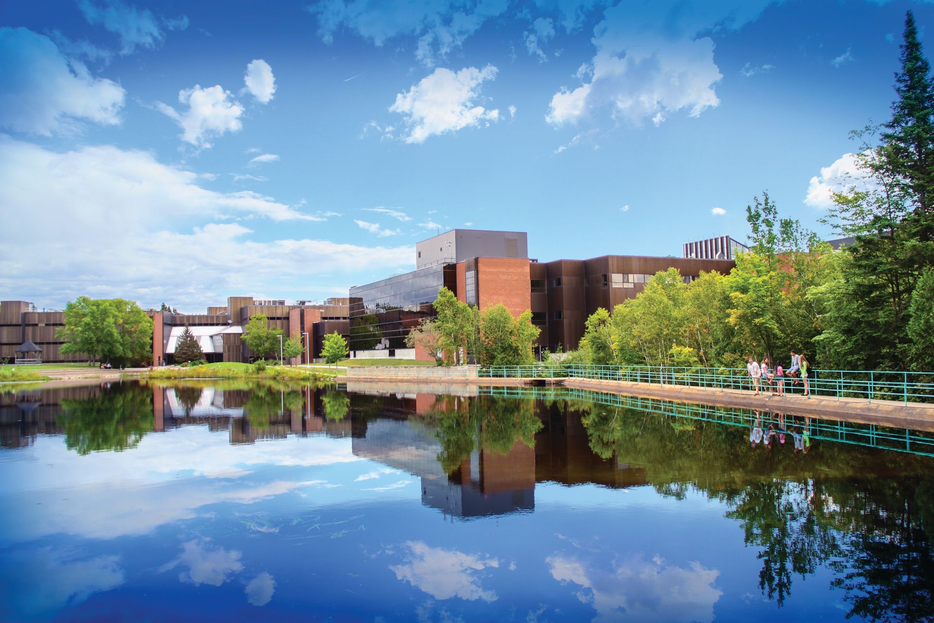 A sunny day view of Nipissing University building complex reflected in calm water, with trees and blue sky.