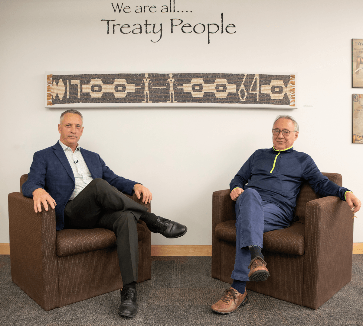 Two men in armchairs, one in a suit, one in casual wear, in an office setting. Sign says