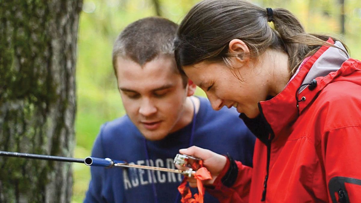 A man and woman in a forest, examining equipment. The woman smiles.