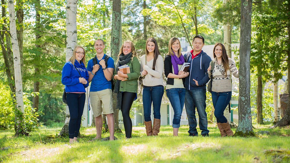 Group of seven people standing in a sunny, grassy area with trees. They wear casual clothes and smile.