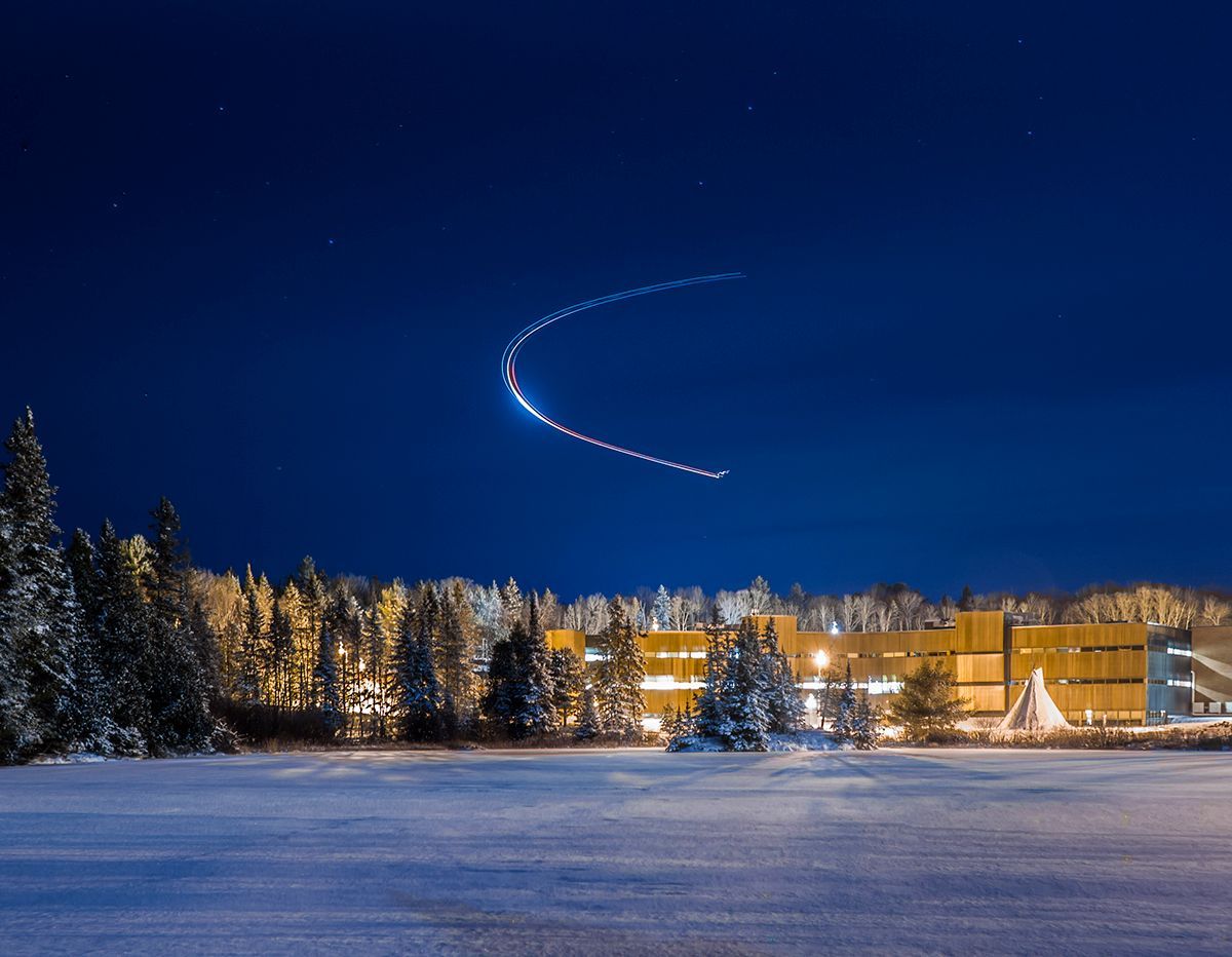 Night scene: airplane light trail arcs across dark blue sky over snowy landscape and lit building.