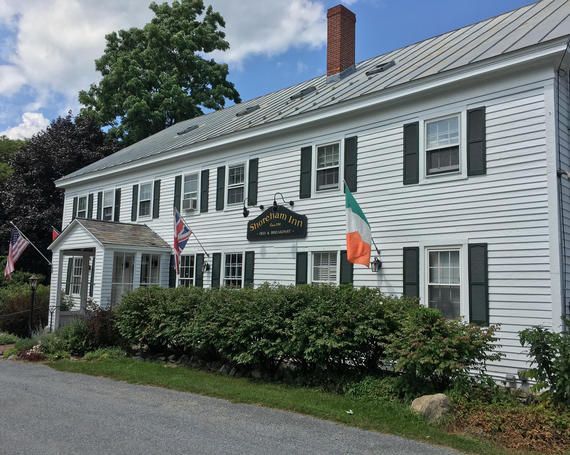 White colonial-style building with shutters, shrubs, and flags beside a paved road