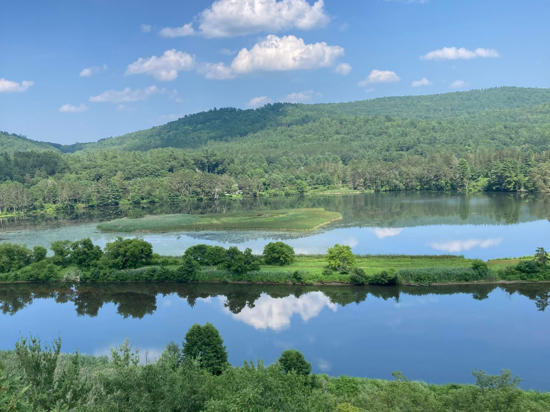 Calm lake reflecting blue sky, green hills, and clouds under a bright sunny landscape.