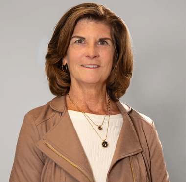A woman with brown hair and a white shirt and brown jacket is smiling for the camera.