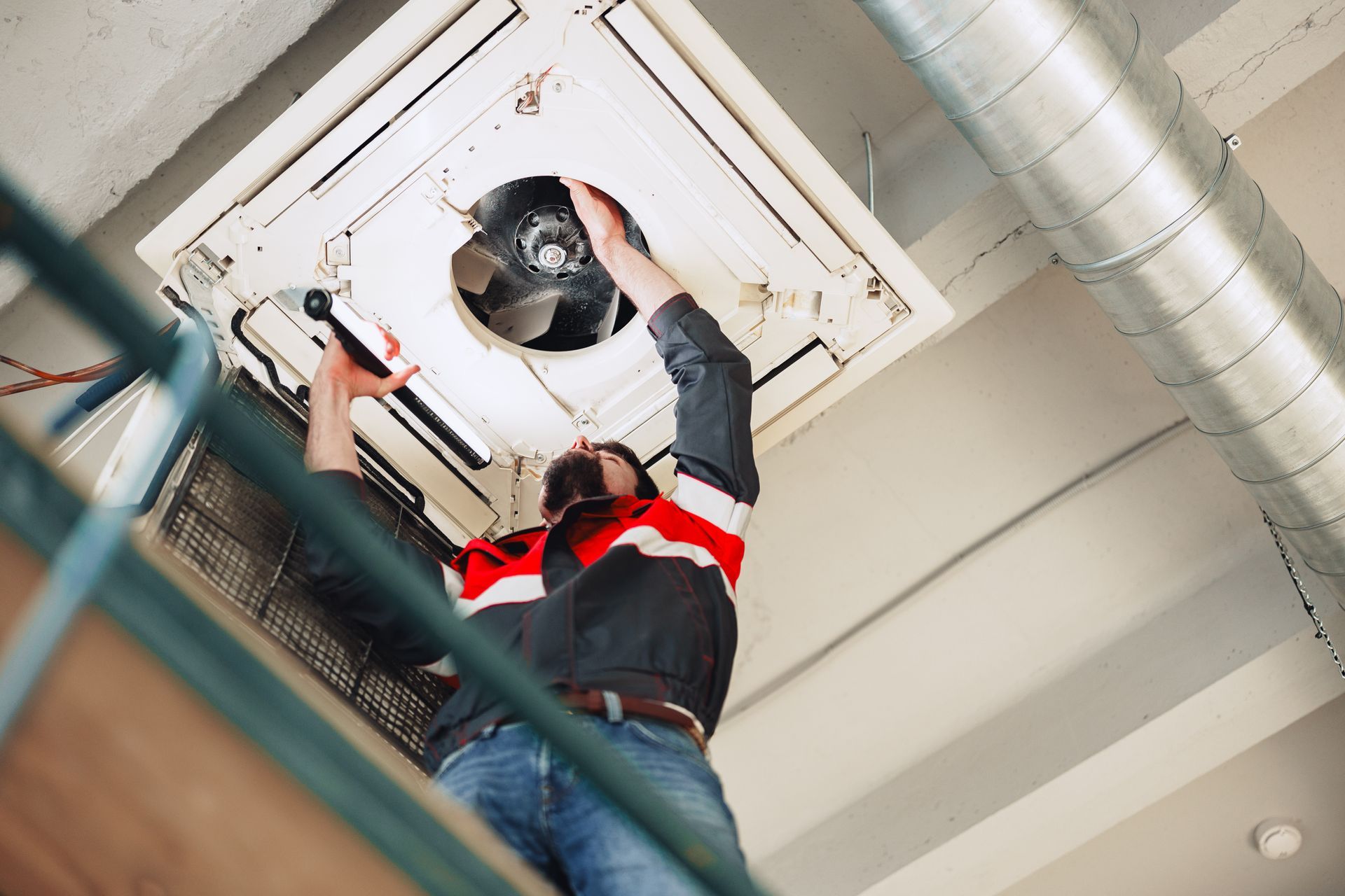 Technician on a ladder inspecting and repairing a ceiling-mounted HVAC unit with exposed fan. Technician on a ladder inspecting and repairing a ceiling-mounted HVAC unit with exposed fan.