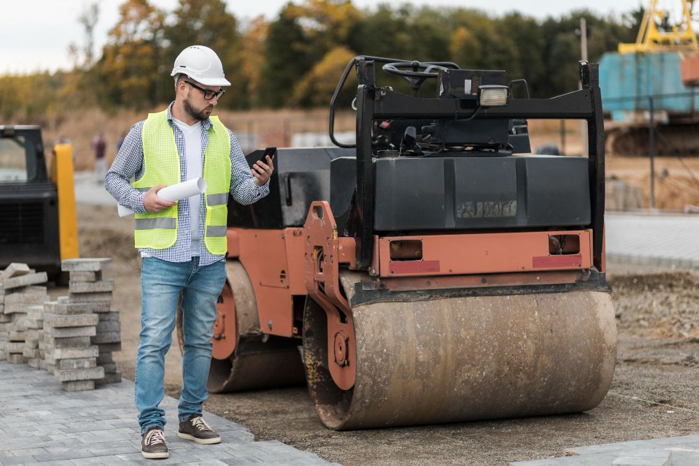 A construction worker is standing next to a roller on a construction site.