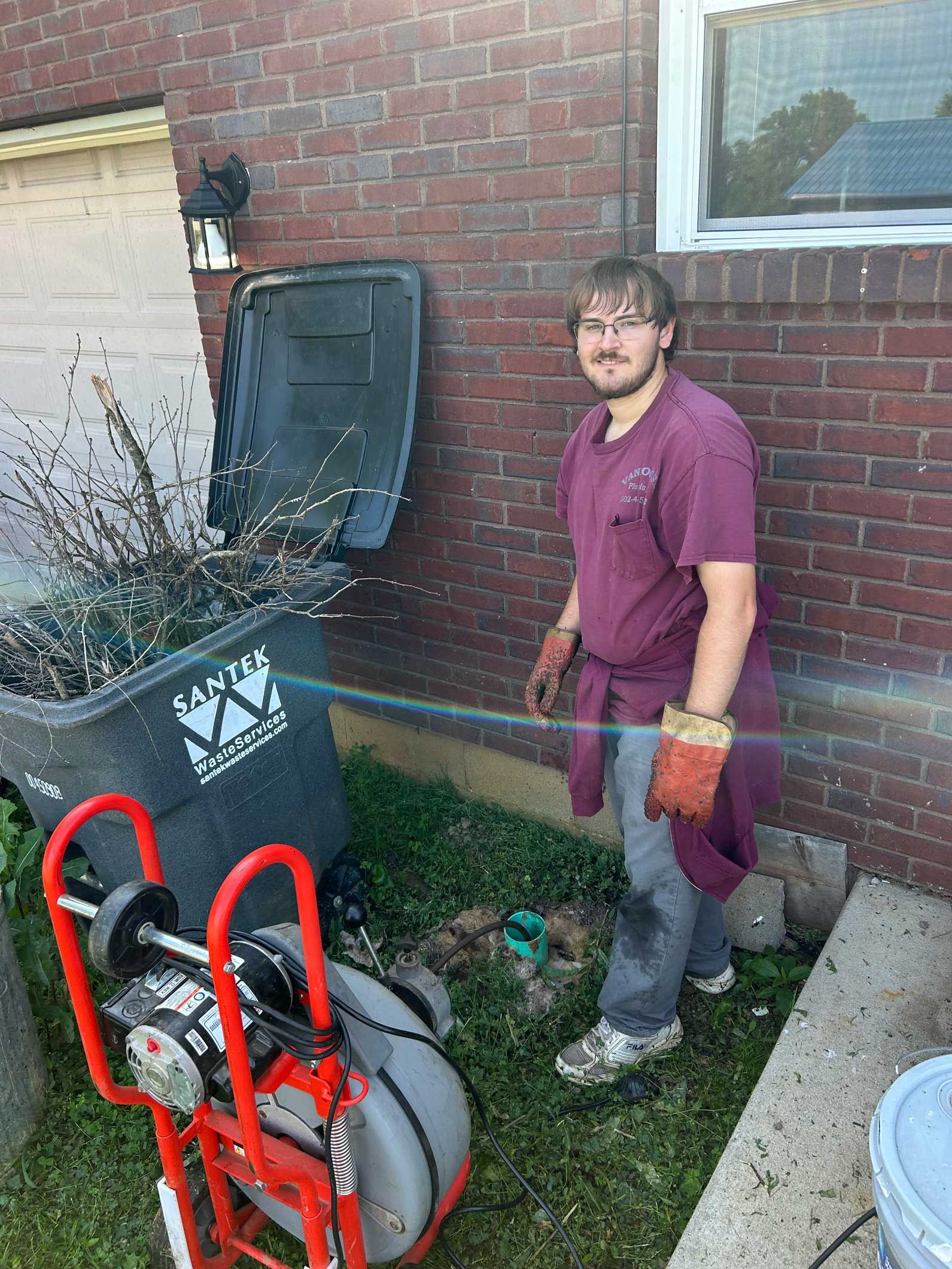 Man in maroon shirt and gloves uses a drain snake machine near a trash can and brick wall.