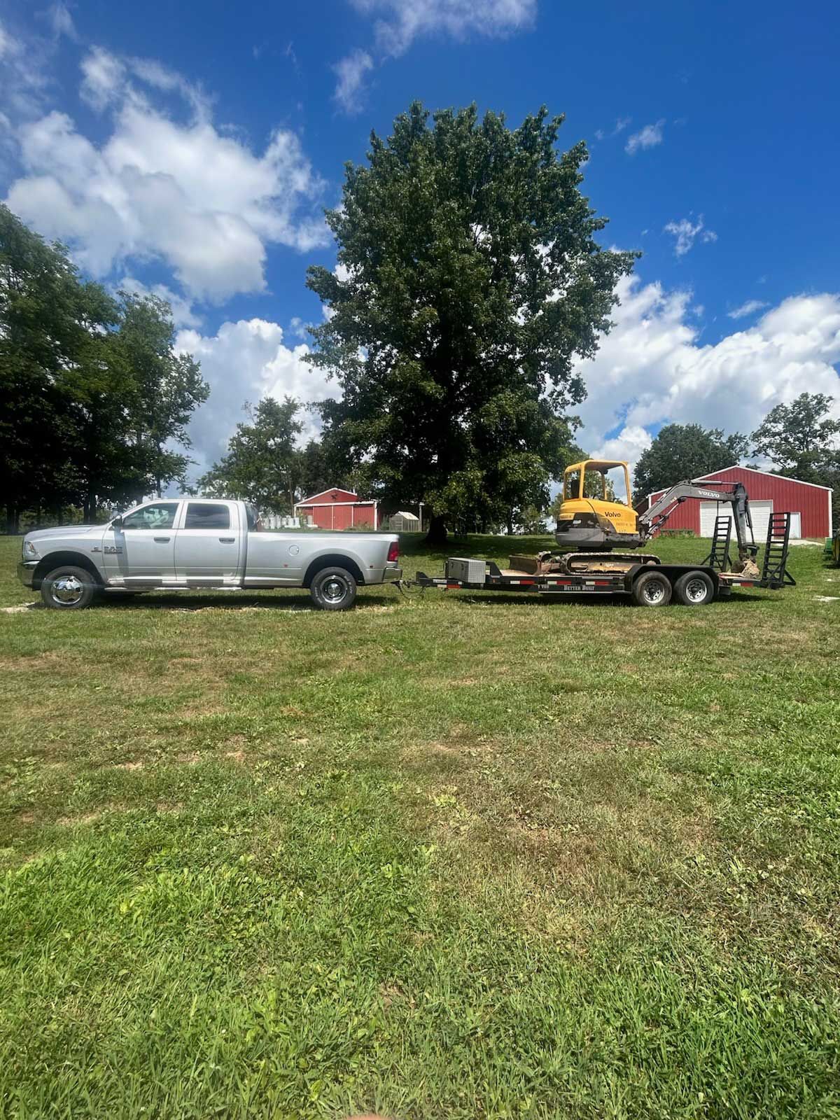 Silver truck pulling a trailer with a yellow excavator on a grassy field, blue sky.