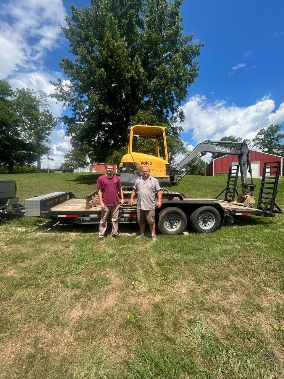 Two men stand in front of a trailer with yellow excavator. Green grass, blue sky, and a red barn in the background.