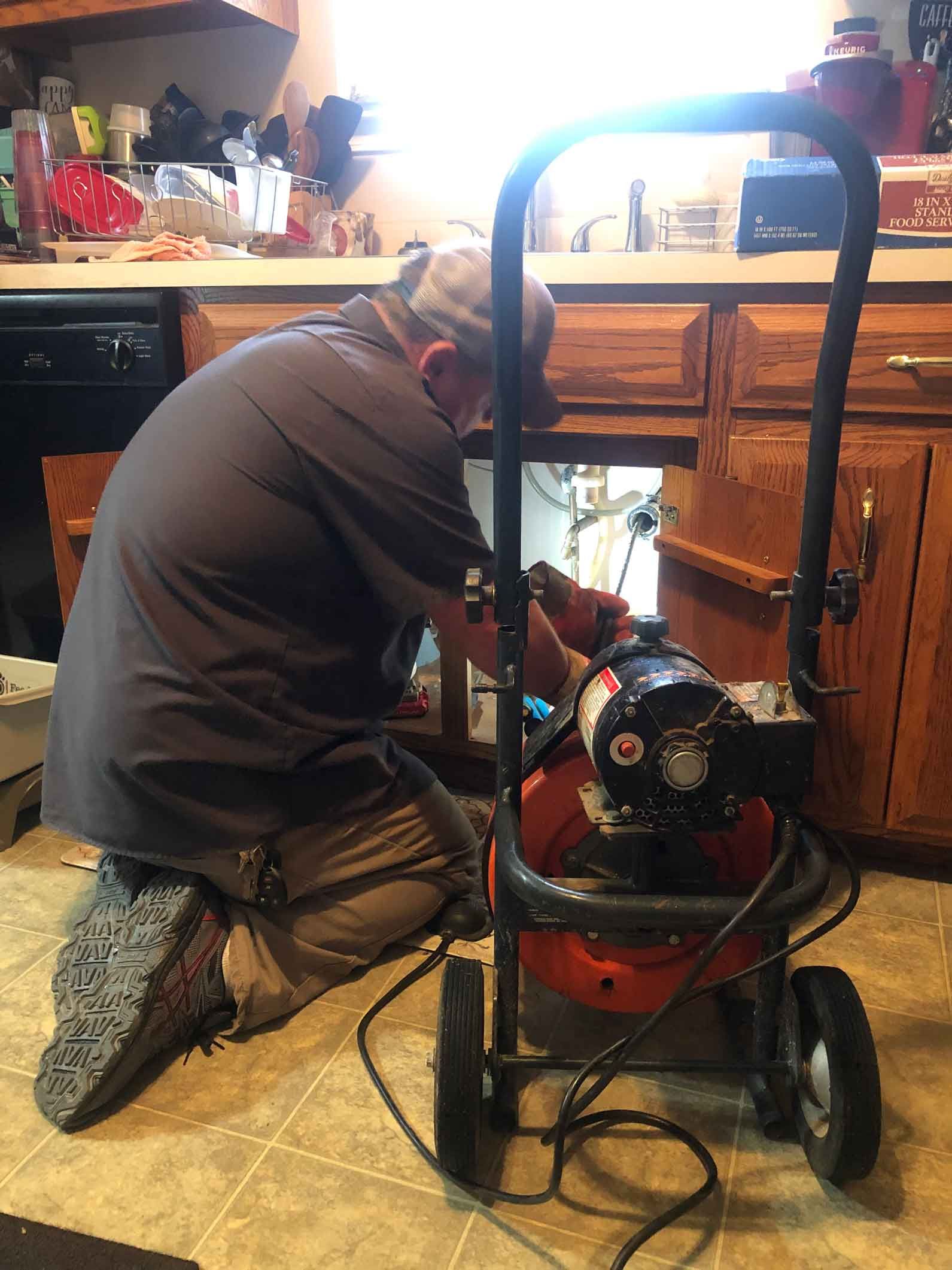 Man kneeling, working on plumbing under kitchen cabinets with a pressure washer nearby.