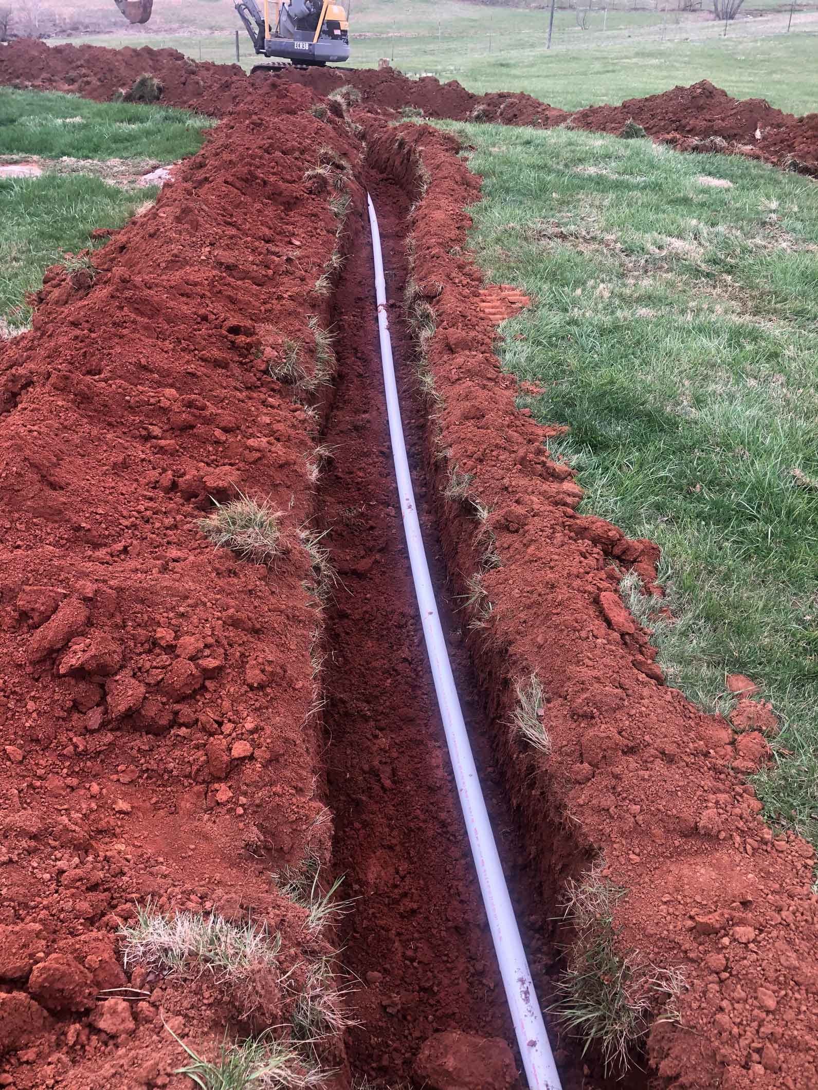 Trench dug in red soil with a white pipe inside, on a green grassy area; excavator in the distance.