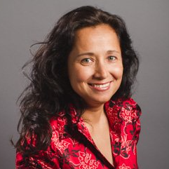 Woman with dark wavy hair smiles, wearing a red patterned jacket against a gray backdrop.