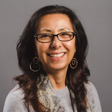 Woman with glasses smiling at camera, wearing earrings and scarf. Gray background.