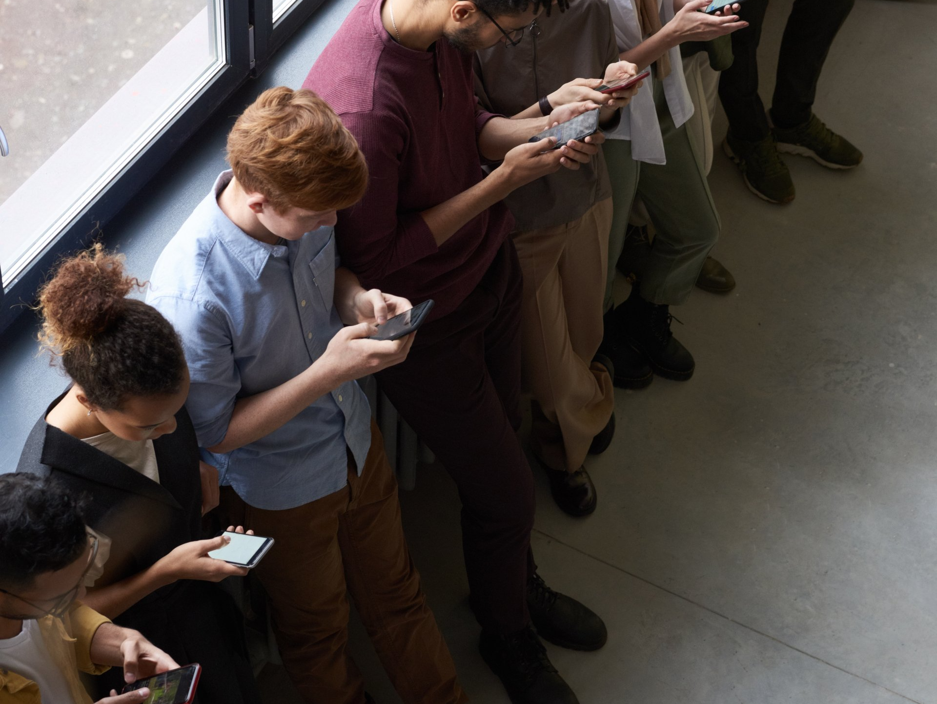 People in a line, looking at smartphones; indoors, near a window.