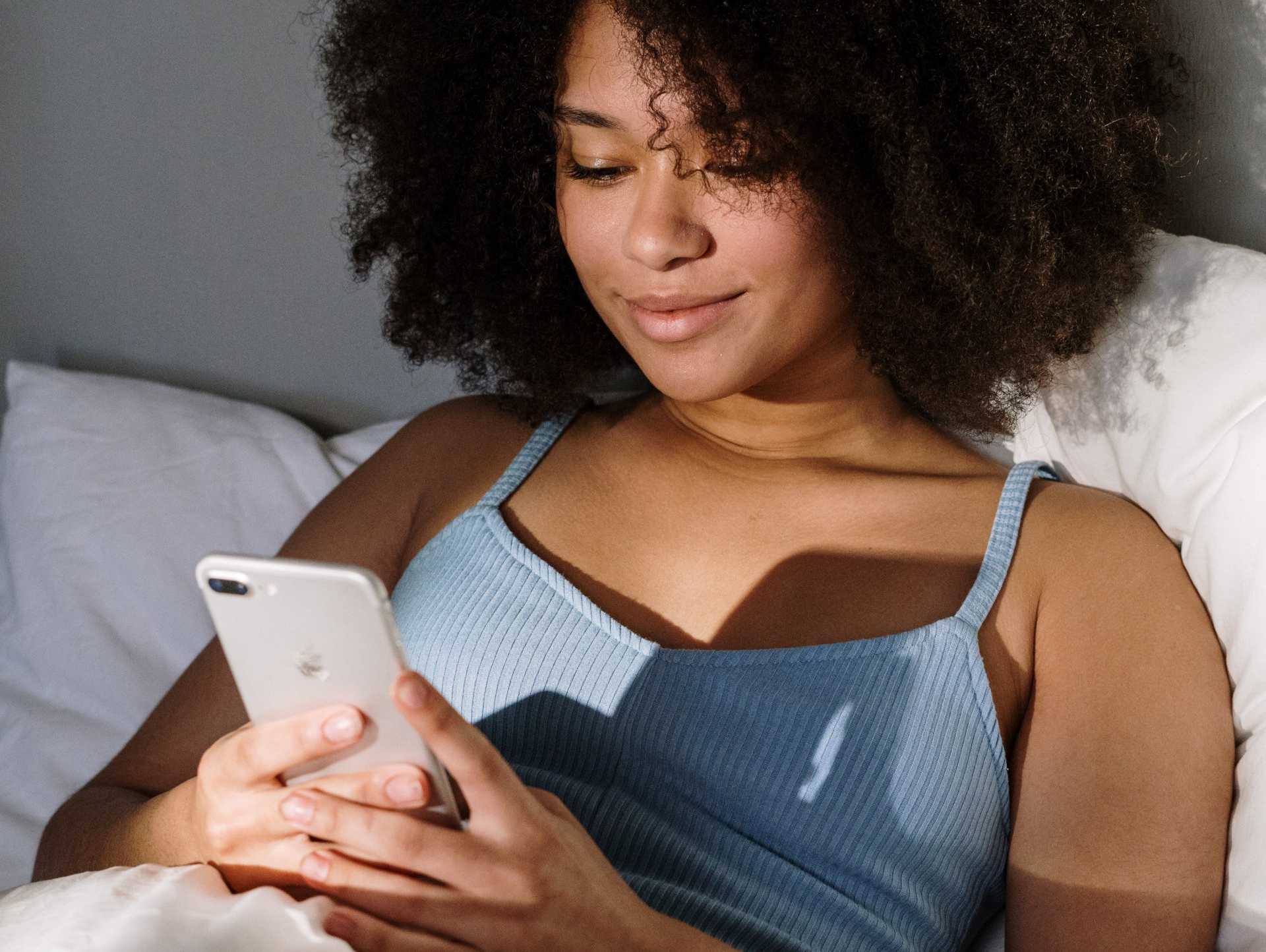 Woman in blue tank top looks at phone while in bed; sunlight illuminates her.