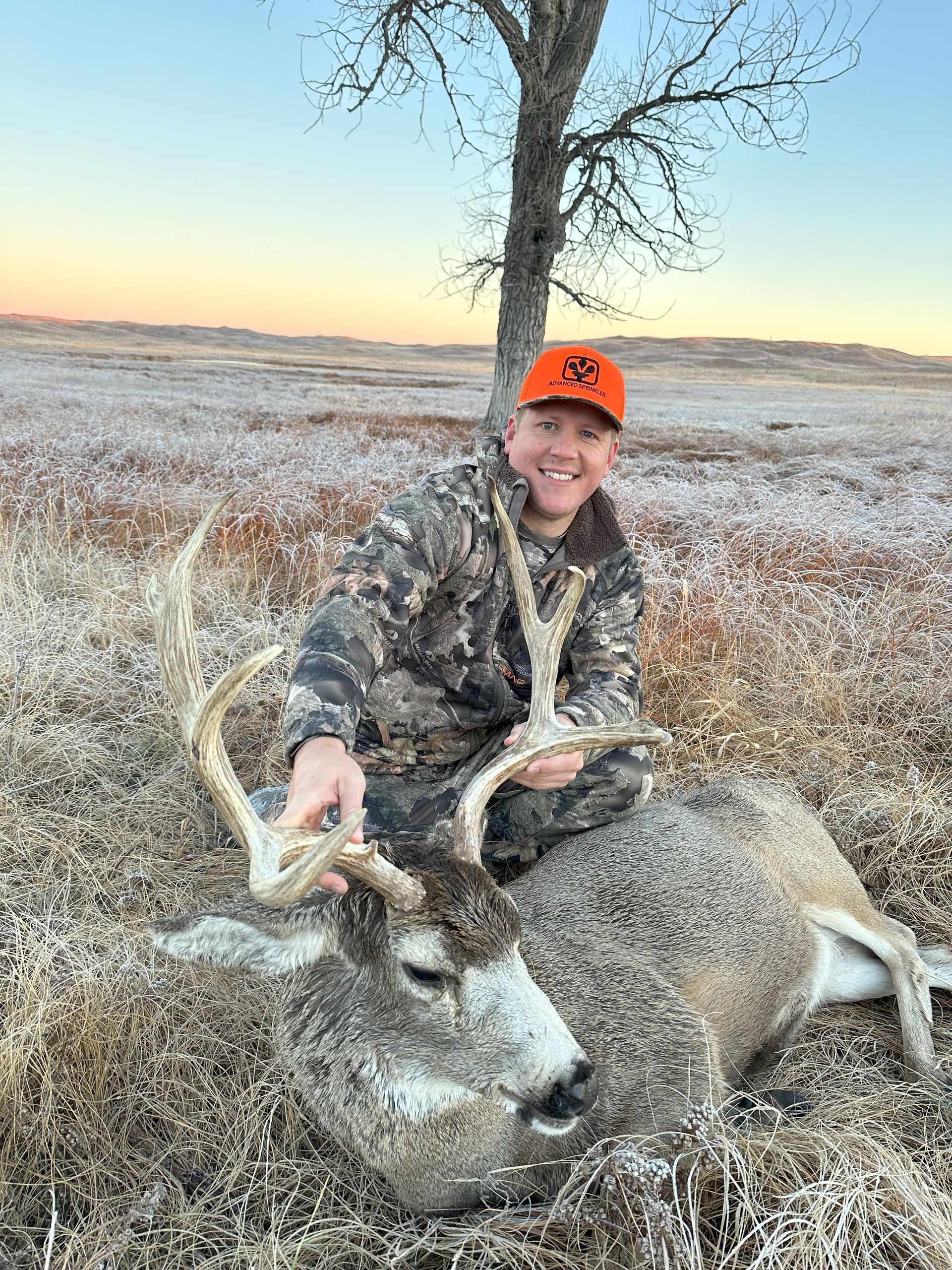 Man in camo and orange hat kneels with a large buck in a field. The man smiles, holding the deer's antlers.