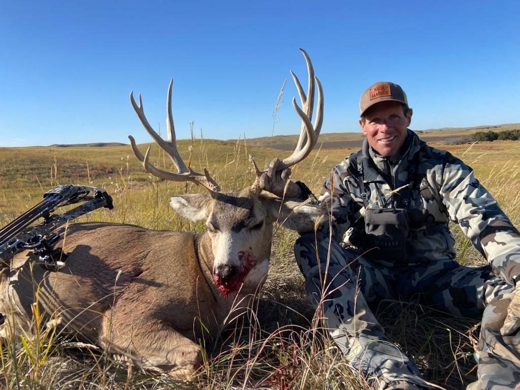 Hunter kneels beside a large buck with a bow, smiling in a grassy field under a blue sky.