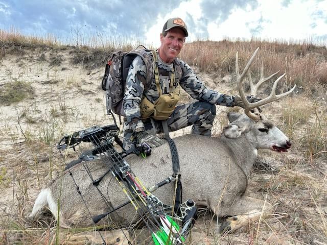 Man kneeling next to a harvested deer, bow and arrow present. Plains setting, camo clothing, smiling.