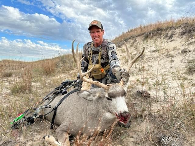 Hunter with deer. Man in camo, smiles, kneels next to buck with bow in grassy field.