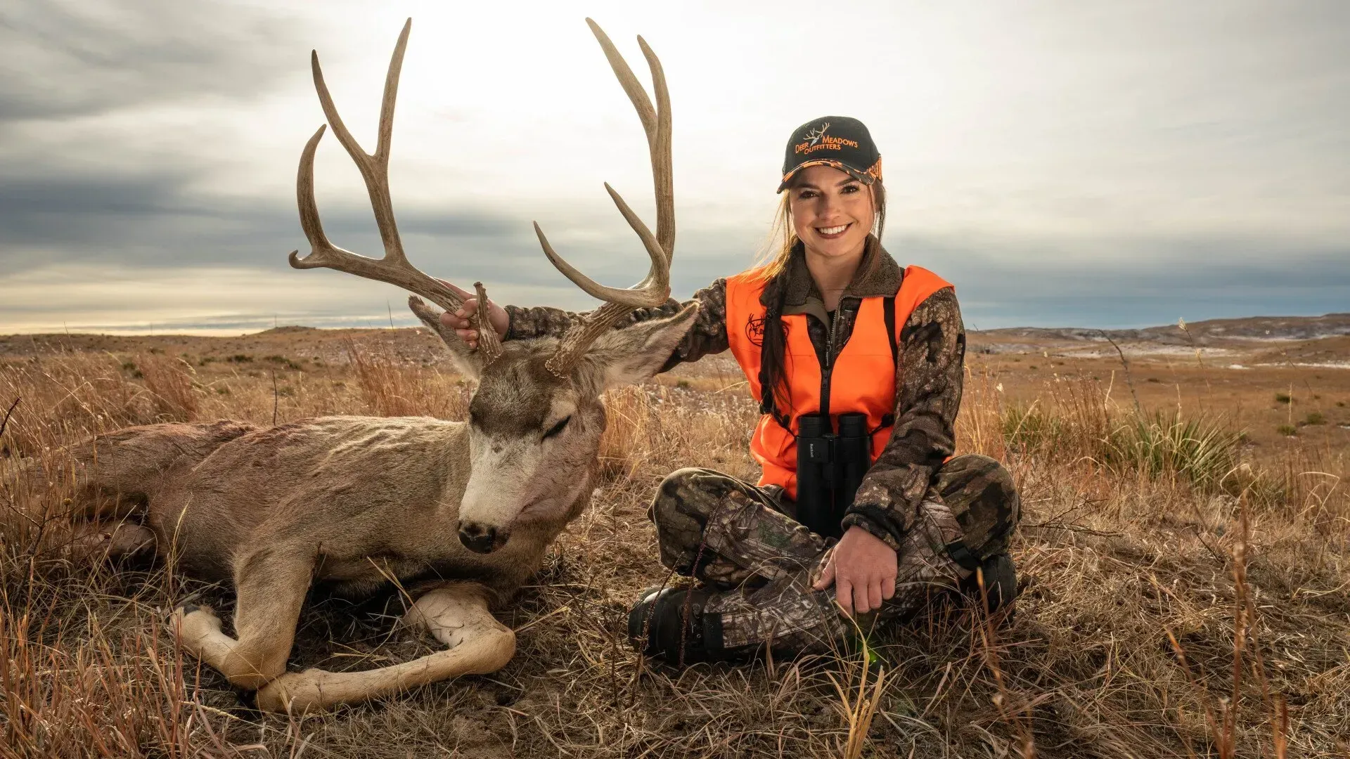 Woman hunter kneels beside a large buck with antlers in a grassy, outdoor setting. She wears camo and orange, smiling.